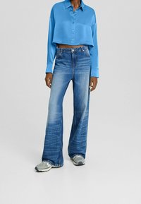 Persona che indossa una camicia corta button-up in satin blu chiaro, jeans a vita alta sbiaditi e larghi di colore blu, e sneaker grigie.