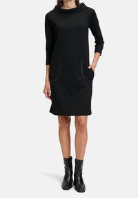 Svart klänning med hög, vikbar krage, trekvartsärmar och två framfickor med en subtil textur. Bärs med svarta ankelskor.