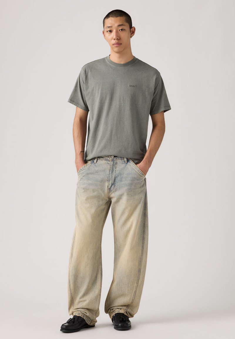 T-shirt grigia a maniche corte con un piccolo logo, abbinata a jeans oversize azzurri chiari. I jeans presentano una sfumatura graduale dal blu al beige. Scarpe nere.