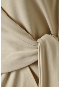 Tissu crème avec une texture lisse, présentant un détail noué et un design drapé. Les coutures et les plis visibles créent de la dimension.