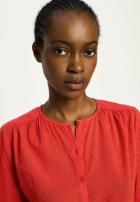 Junge Frau mit geflochtenem Haar, die eine strukturierte rote Bluse mit Knöpfen vor einem schlichten hellen Hintergrund trägt.