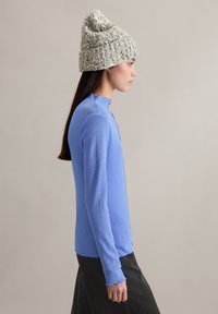 Langärmeliges, hellblaues, geripptes Oberteil mit hohem, gewelltem Kragen. Das Modell trägt eine strukturierte, hellgraue, gestrickte Mütze.