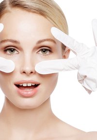 Modella con pelle chiara e capelli castano chiaro, indossa guanti bianchi e posa con le mani che formano una forma a V sul volto su uno sfondo bianco.