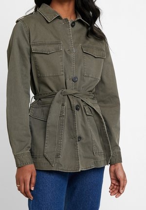 Femme portant une veste boutonnée vert olive avec poches poitrine et hanches, nouée à la taille, associée à un jean bleu.