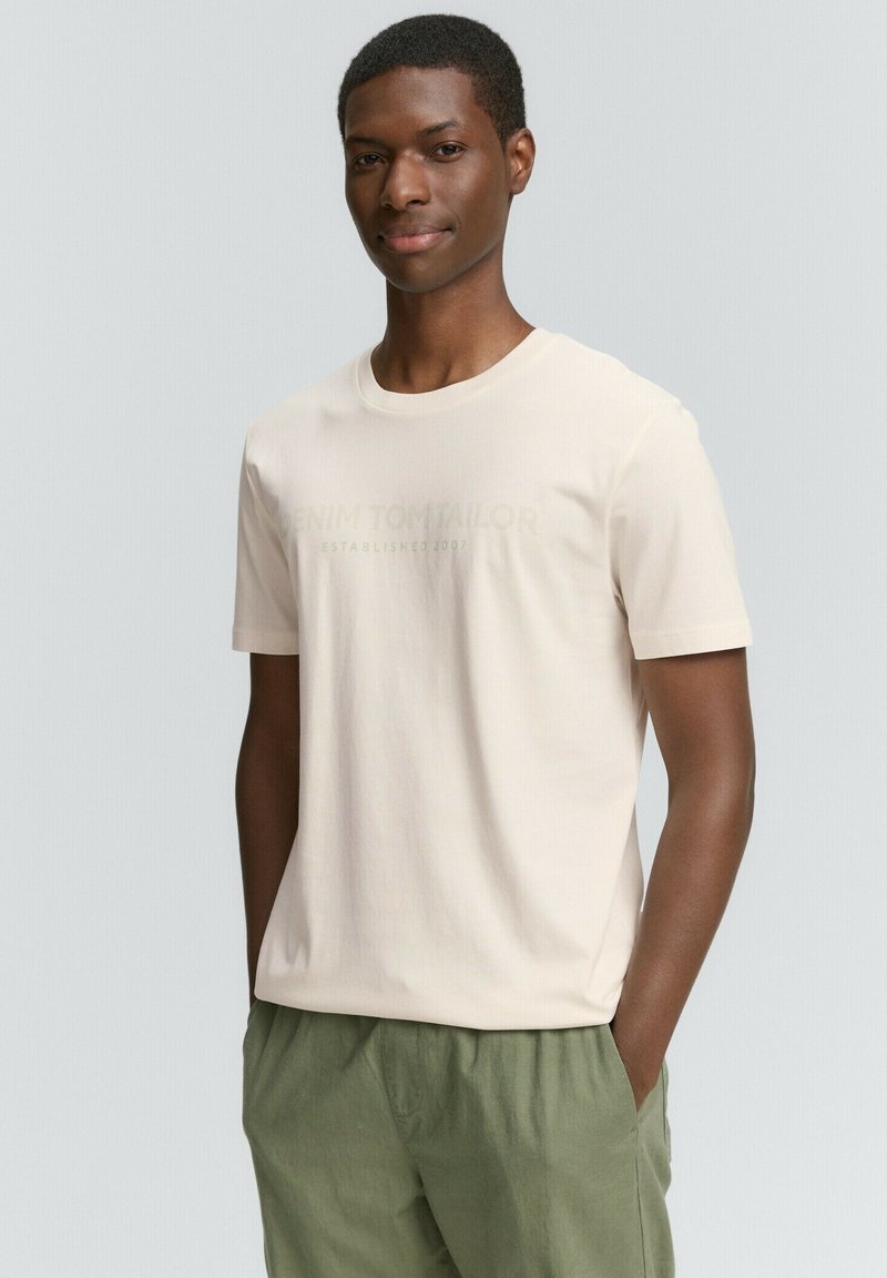 Lys beige bomulds t-shirt med rund hals og korte ærmer, der har et diskret, prægede logo i grøn. Sæt sammen med olivengrønne shorts.