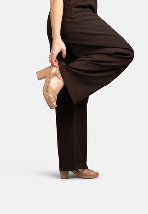 Persoon in bruine broek die een beige suède sandaal met blokhak aanpast op een geheven voet tegen een egale lichte achtergrond.