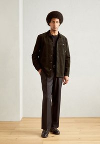 Chaqueta de pana verde oscuro con cierre de botón, bolsillo en el pecho, camiseta negra, pantalones negros ajustados y zapatos negros con detalles mínimos.