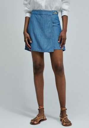 Mujer con una falda vaquera azul estilo envolvente con una pequeña hebilla de metal, combinada con una camisa blanca y sandalias marrón claro con pulsera en el tobillo.
