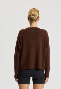 Hnedý pletený sveter s rebrovanou textúrou a okrúhlym výstrihom, na zadnej strane s čiernou nálepkou. Kombinovaný s čiernymi šortkami.