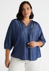 Blusa in denim blu, leggera, con scollatura a V, dettagli con cordoncino e maniche a tre quarti con polsini arricciati. Tessuto morbido.