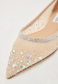 Beige litasuulehke imeliked seena koos võrkkate üksikasjadega, mis on kaunistatud selgete ja iiri rhinestone'idega ning sädeleva kristallriba avause ääres.