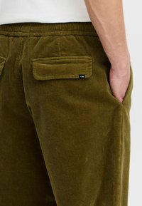 Olive green corduroy pants featuring a soft texture, elastic waistband, and back pockets with flaps. Small brand tag on pocket.