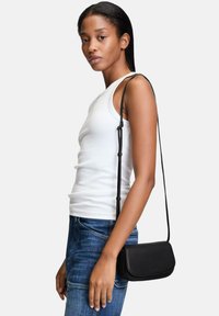 Schwarze Leder-Umhängetasche mit glatter Textur, rechteckiger Form und langem verstellbarem Tragegurt, getragen zu einem weißen Tanktop und blauen Jeans.