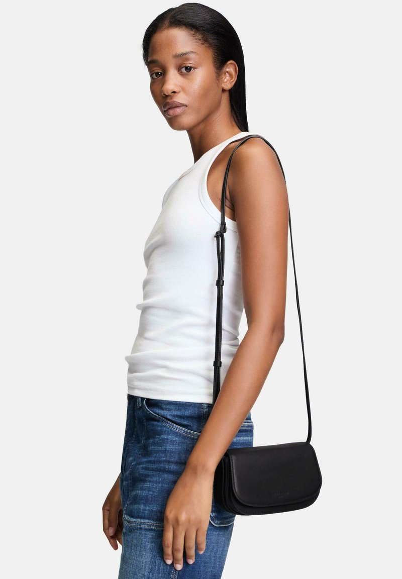 Schwarze Leder-Umhängetasche mit glatter Textur, rechteckiger Form und langem verstellbarem Tragegurt, getragen zu einem weißen Tanktop und blauen Jeans.