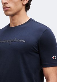 Άνδρας που φοράει μπλε ναυτικό μπλουζάκι Champion με ανυψωμένο λογότυπο στο στήθος και κεντημένο λογότυπο της μάρκας στο αριστερό μανίκι.