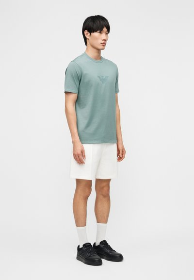 Νέος άνδρας όρθιος, φοράει ανοιχτοπράσινο T-shirt με λογότυπο, άσπρο σορτς, άσπρες κάλτσες και μαύρα αθλητικά παπούτσια σε απλό λευκό φόντο.