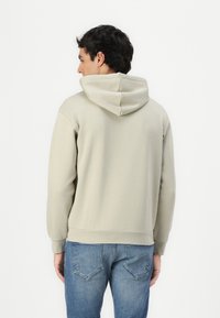 Sweatshirt com capuz em tecido cinza claro, apresentando um corte relaxado, punhos e bainha canelados, combinado com jeans de denim azul. Detalhes de costura visíveis.