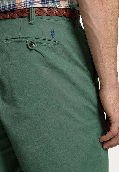 Pantaloni din bumbac verde, cu închidere frontală cu nasture, buzunare laterale și un logo subtil brodat. Purtați cu o curea din piele împletită maro.