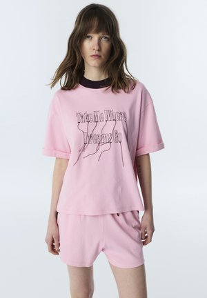 Jonge vrouw met schouderlang haar die een roze set met shirt en korte broek draagt, met de tekst "Take Me Where Dreams Go" op het shirt.