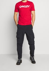 Czerwona koszulka z krótkim rękawem z białym logo "OAKLEY", zestawiona z czarnymi spodniami cargo z wieloma przednimi kieszeniami i gładką fakturą.