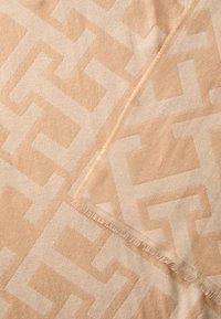 Beige woven blanket with a geometric pattern featuring interlocking shapes, finished with fringe edges for texture and detail.