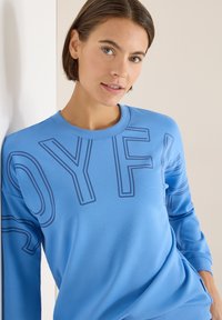 Jasnoniebieska bluza wykonana z miękkiego materiału, z dużymi granatowymi literami "O", "Y", "F" na przodzie. Okrągły dekolt i długie rękawy.