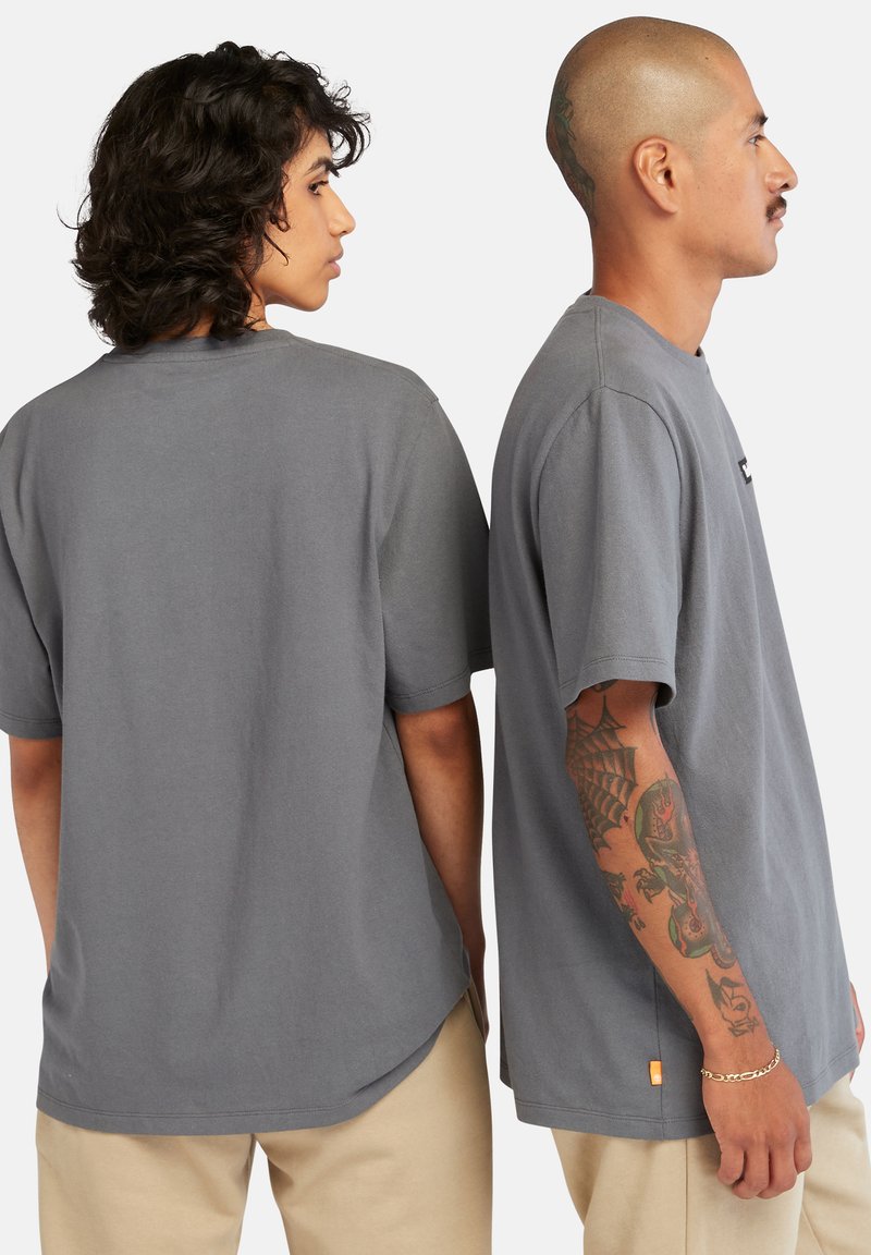 T-shirt in cotone grigio con maniche corte e vestibilità comoda, caratterizzata da un piccolo logo sul petto e un'etichetta sul lato, abbinata a pantaloni beige.