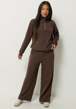 Donna con capelli lunghi e scuri che indossa una felpa marrone con zip e collo alto e pantaloni ampi abbinati con scarpe da ginnastica bianche.
