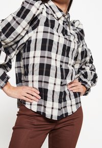 Chemise  à carreaux en noir, blanc et marron, présentant un design à boutons, des manches bouffantes et un col classique. Texture de tissu lisse.