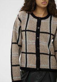 Bežs adīts kardigāns ar melnu un gaiši pelēku svītrainu rakstu, ar melnām pogām priekšpusē, ribotu kakla izgriezumu un garām piedurknēm.