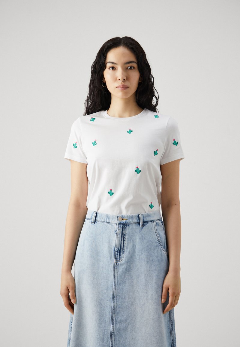 Camiseta de algodón blanca con bordado de pequeño cactus verde y flores rosas, combinada con una falda de mezclilla azul claro. Diseño simple de cuello redondo.