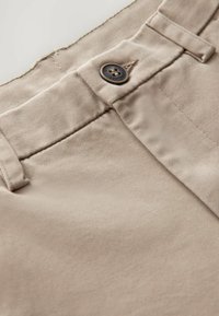 Pantaloni in cotone beige con una texture liscia, tasche laterali e un bottone marrone in chiusura in vita. Dettagli di cuciture fini visibili.