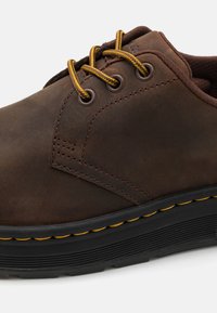 Rjave usnjene čevlje z gladko teksturo, ki imajo rumene vezalke, okrogla očesca in debel črn podplat z rumenim dekorativnim šivom.