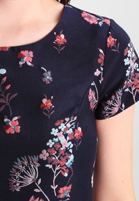 Námořnická modrá blůza s krátkým rukávem s květinovým vzorem v červené, růžové, bílé a tyrkysové barvě na světlém pozadí.