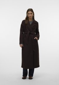 Cappotto lungo marrone con cintura, rever a lancia e tasche laterali, abbinato a un top grigio e jeans scuri. Tessuto liscio, vestibilità su misura.