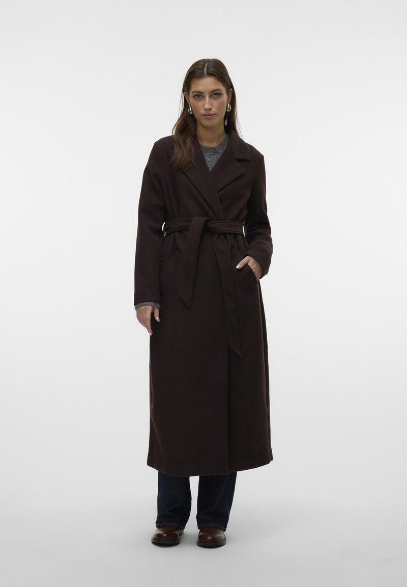 Cappotto lungo marrone con cintura, rever a lancia e tasche laterali, abbinato a un top grigio e jeans scuri. Tessuto liscio, vestibilità su misura.