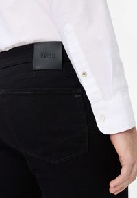 Jeans neri con una texture liscia, dotati di una toppa in pelle sul retro e cuciture dettagliate. Camicia bianca con polsini abbottonati visibili.