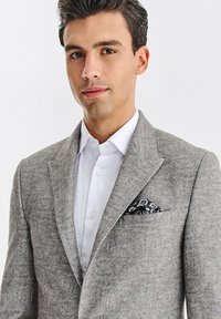 Szary wełniany żakiet z klapami notch, noszony na białej koszuli. Posiada czarno-niebieski wzorzysty poszetkę w kieszeni piersiowej.