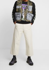 Maskovací vesta se šedými skvrnami, černé dlouhé tričko s grafikou, béžové kalhoty s širokými nohavicemi a  černé tenisky. Viditelné texturované materiály.