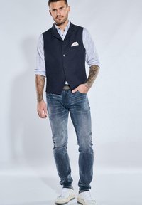 Marineblaues Weste mit einer Tasche, getragen über einem hellblau gestreiften Hemd. Hellblaue Jeans und weiße Sneakers vervollständigen das Outfit.