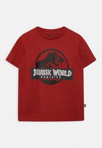 Röd bomullströja med en svart dinosauriegrafik och texten "JURASSIC WORLD DOMINION" på framsidan. Korta ärmar, rundad halsdesign.