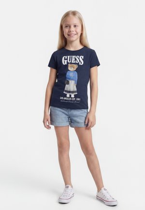 Smilende pige med langt blondt hår iført en marineblå Guess T-shirt med bjørneprint, denimshorts og hvide sneakers, stående foran en hvid baggrund.
