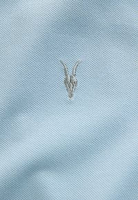Světle modrá texturovaná látka s malým stříbrným vyšívaným designem lebky kozy ve středu.