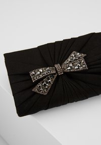 Pochette en tissu noir avec un design plissé et un nœud décoratif central orné de strass noirs et argentés.