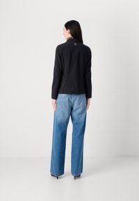 Schwarze Jacke mit langen Ärmeln, Logo-Detail auf dem Rücken, kombiniert mit weit geschnittenen hellblauen Jeans und schwarzen spitzen Heels. Einfaches Design, glatte Textur.
