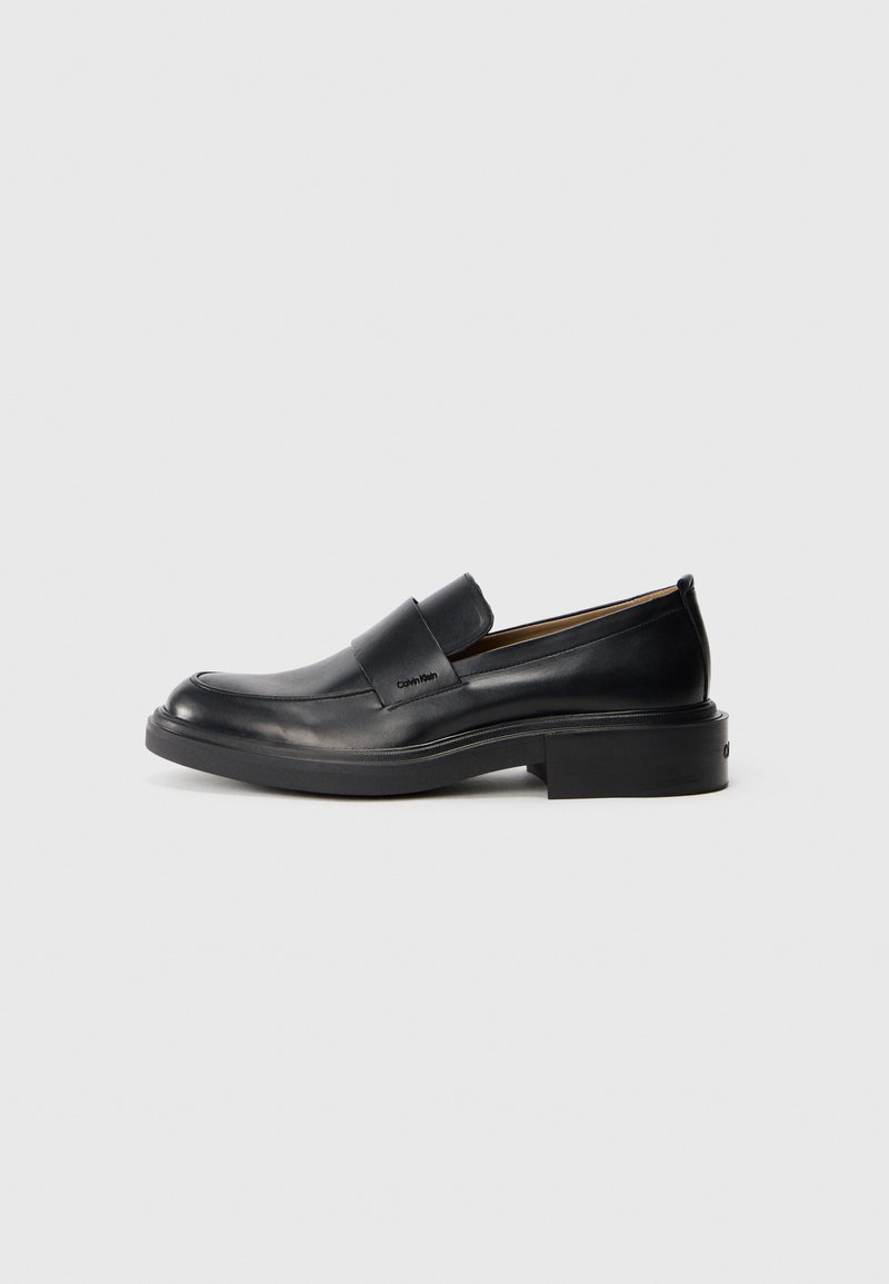 Pantofle negre din piele pentru bărbați, cu toc mic și un logo subtil Calvin Klein pe lateral, prezentate în profil pe un fond alb.