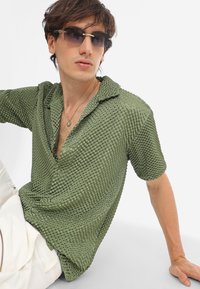 Olijfgroene, gestructureerde short-sleeve shirt met knoopsluiting en een kraag, gecombineerd met donkere zonnebril en een hanger ketting.