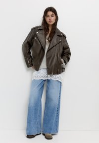 Junge Frau trägt eine übergroße braune Lederjacke, einen grauen Pullover, ein top mit Spitzenaufschlägen, weite blaue Jeans und braune Stiefel, steht vor einer weißen Wand.