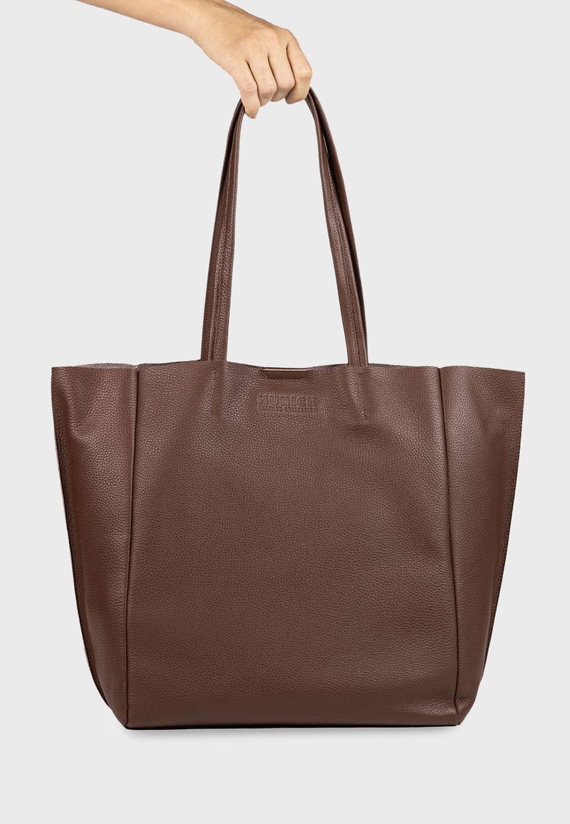 Main tenant un sac fourre-tout en cuir marron texturé avec deux longues poignées sur un fond clair uni.