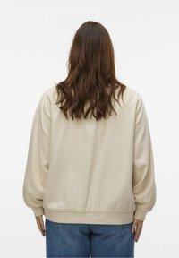 Ljusbeige bomberjacka med puffärm, ribbat midjeband och slät yta, kombinerad med blå denimjeans. Bakvy av outfiten.